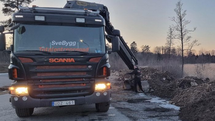 Svart lastbil, Scania-text i orange färg, SveBygg Totalentreprenad står på lastbilens framruta, på flaket finns en grävskopa